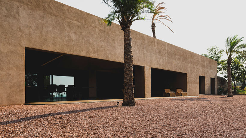 Edificio moderno dal design minimale con grandi aperture, pareti materiche color tortora e dettagli esotici